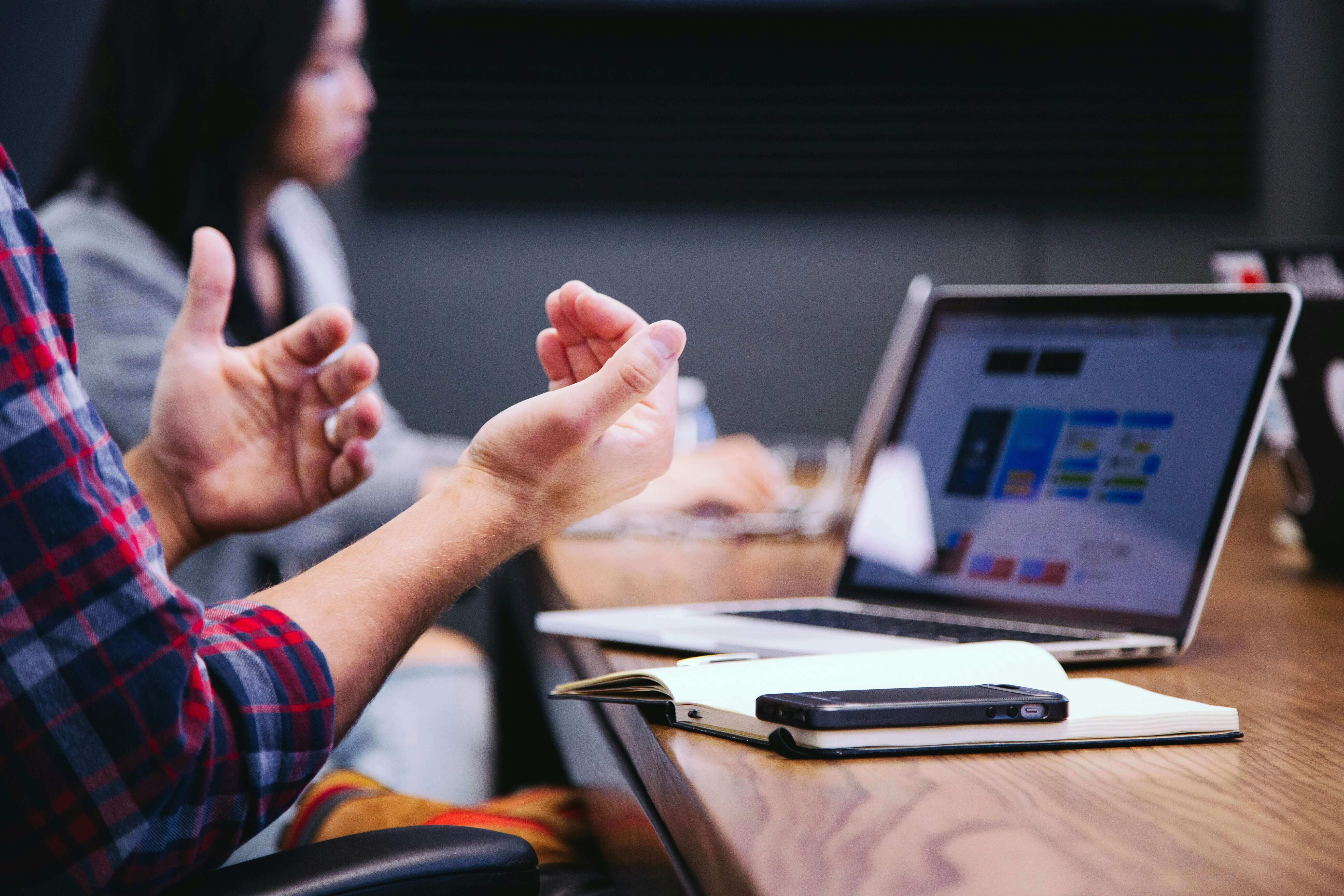 A person gesturing with their hands during a discussion at a table with a laptop displaying a presentation