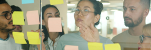 A group of people in an office setting, collaborating over post it notes stuck to a glass partition.