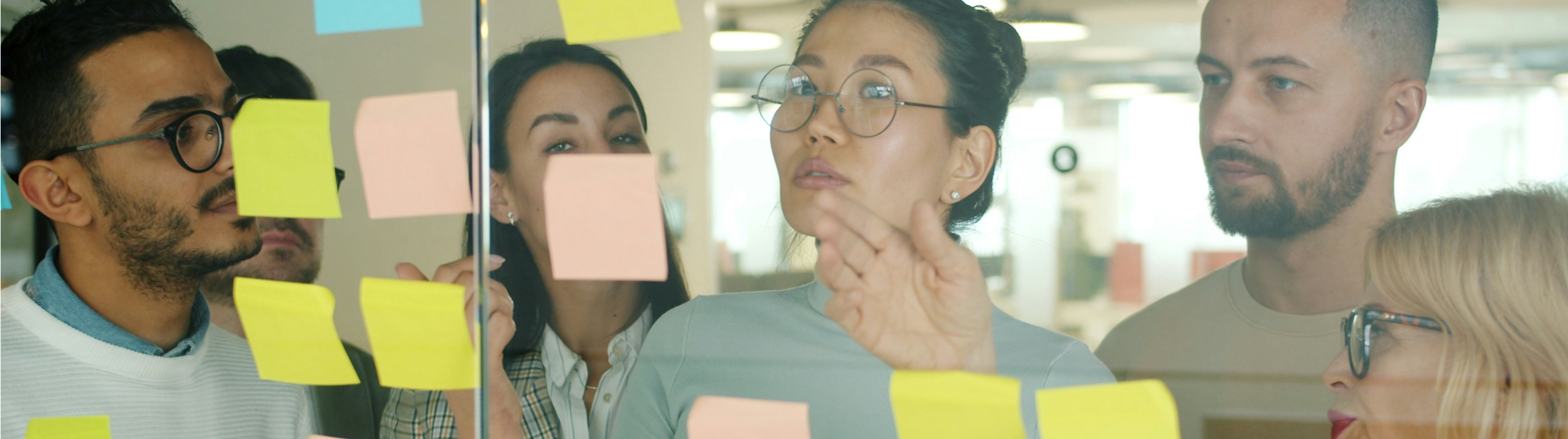 A group of people in an office setting, collaborating over post it notes stuck to a glass partition.