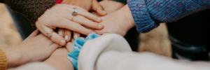 Close up of people's arms wearing colourful sweaters, pressing their hands together in a circle.