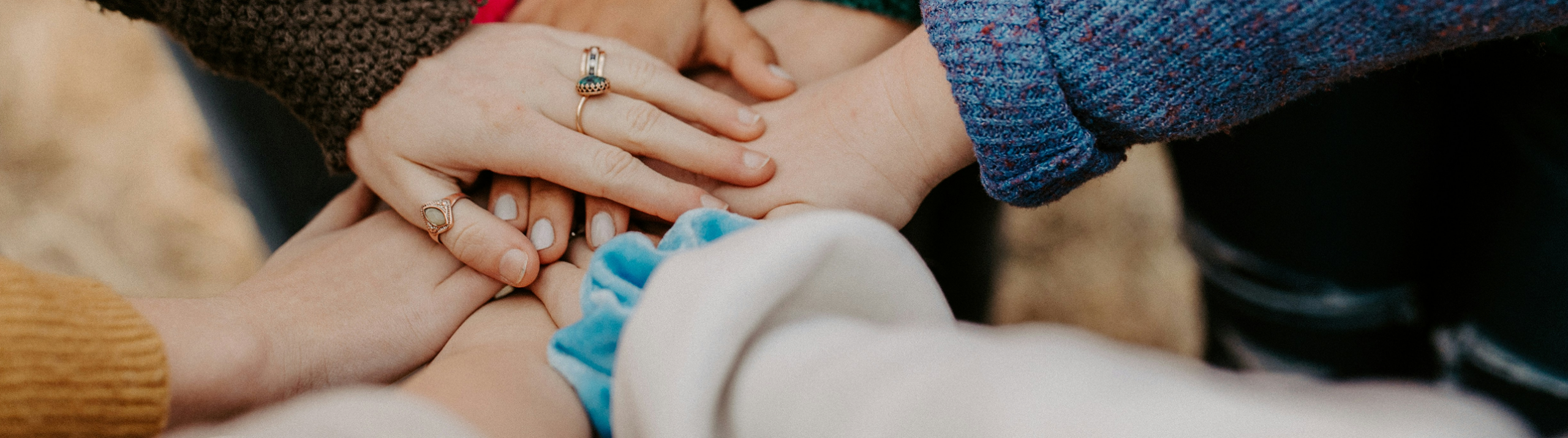 Close up of people's arms wearing colourful sweaters, pressing their hands together in a circle.
