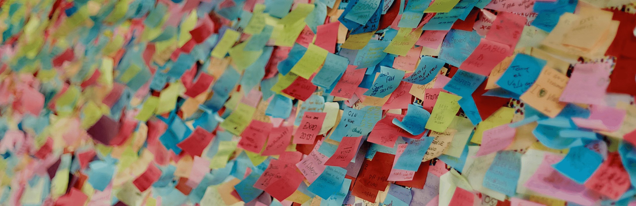 Colorful wall covered with numerous handwritten sticky notes in blue, pink, yellow, and red tones, forming a dense collage of messages and names at what appears to be an event or exhibition space.